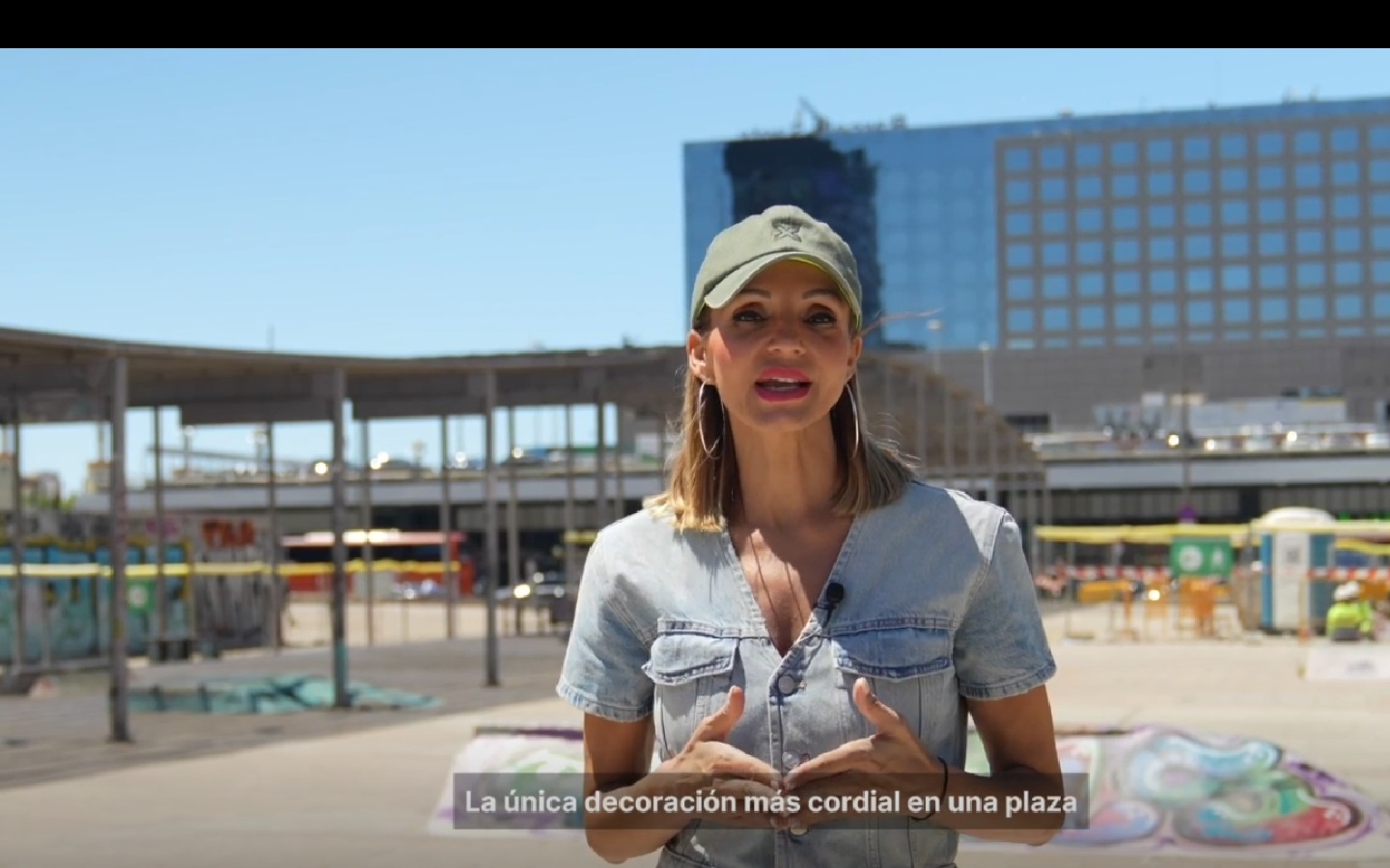 Reportera ante la plaza de la Estación de Sants, ante varios graffitis. En la parte inferior de la imagen puede verse el subtítulo de lo que dice ella en el momento, que es: "La única decoración más cordial en una plaza"Reportera ante la plaza de la Estación de Sants, ante varios graffitis. En la parte inferior de la imagen puede verse el subtítulo de lo que dice ella en el momento, que es "La única decoración más cordial en una plaza".